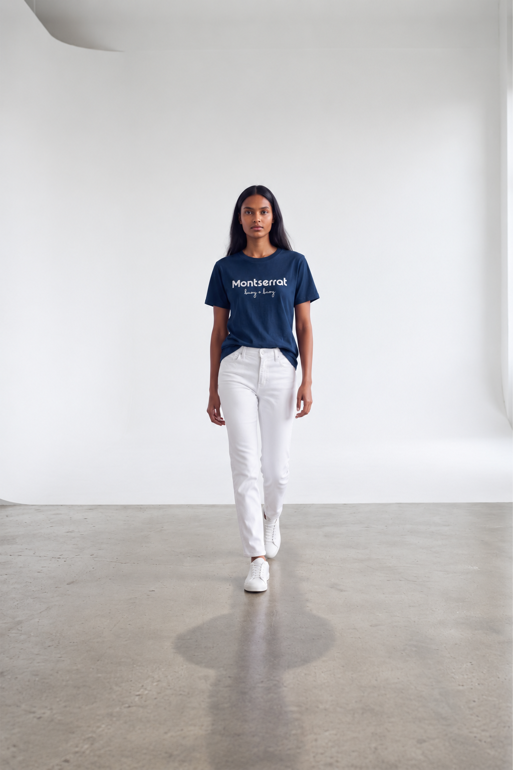A woman walks toward the camera in a minimalist studio setting, wearing a navy blue Montserrat graphic t-shirt, white pants, and white sneakers.