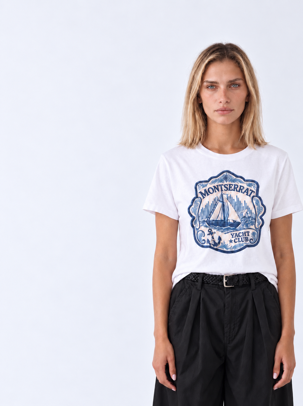 A woman modeling a white short-sleeve t-shirt with a blue vintage-style 'Montserrat Yacht Club' graphic on the front, paired with black trousers, against a plain light gray background.