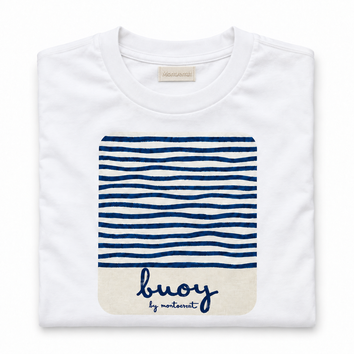 A folded white crewneck t-shirt by Montserrat, featuring a navy blue and white striped graphic on the front with a cream-coloured bottom section containing the brand name 'buoy by Montserrat' in navy script.